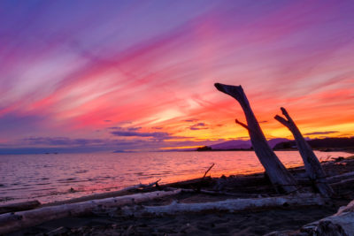 Fiery pink, purple, and orange sunset at Iona Beach framed by driftwood in foreground and North Shore Mountains in background