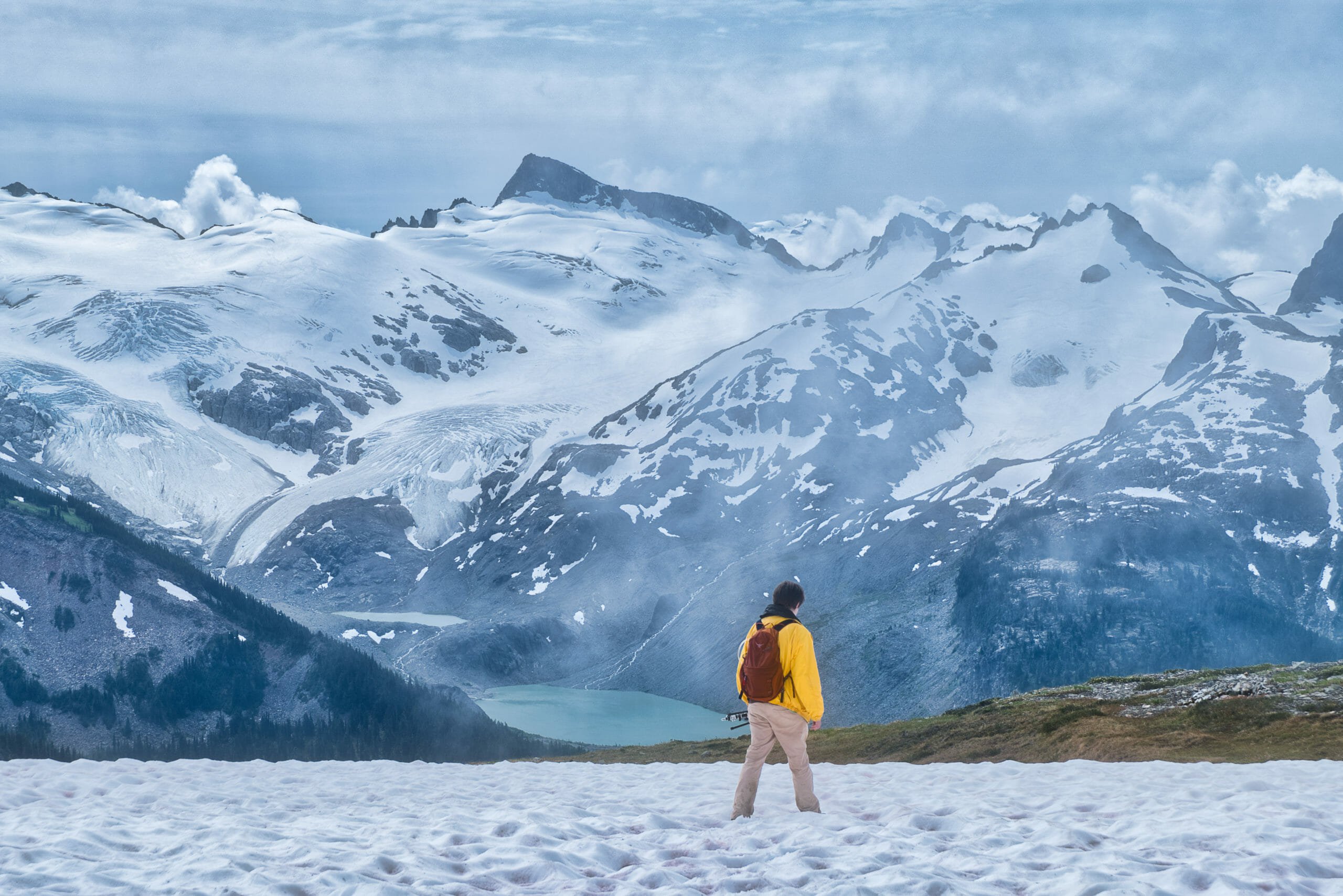 Male hiker treks through snowfield toward alpine lake with snowy mountain ranges in Garibaldi Provincial Park on cloudy day