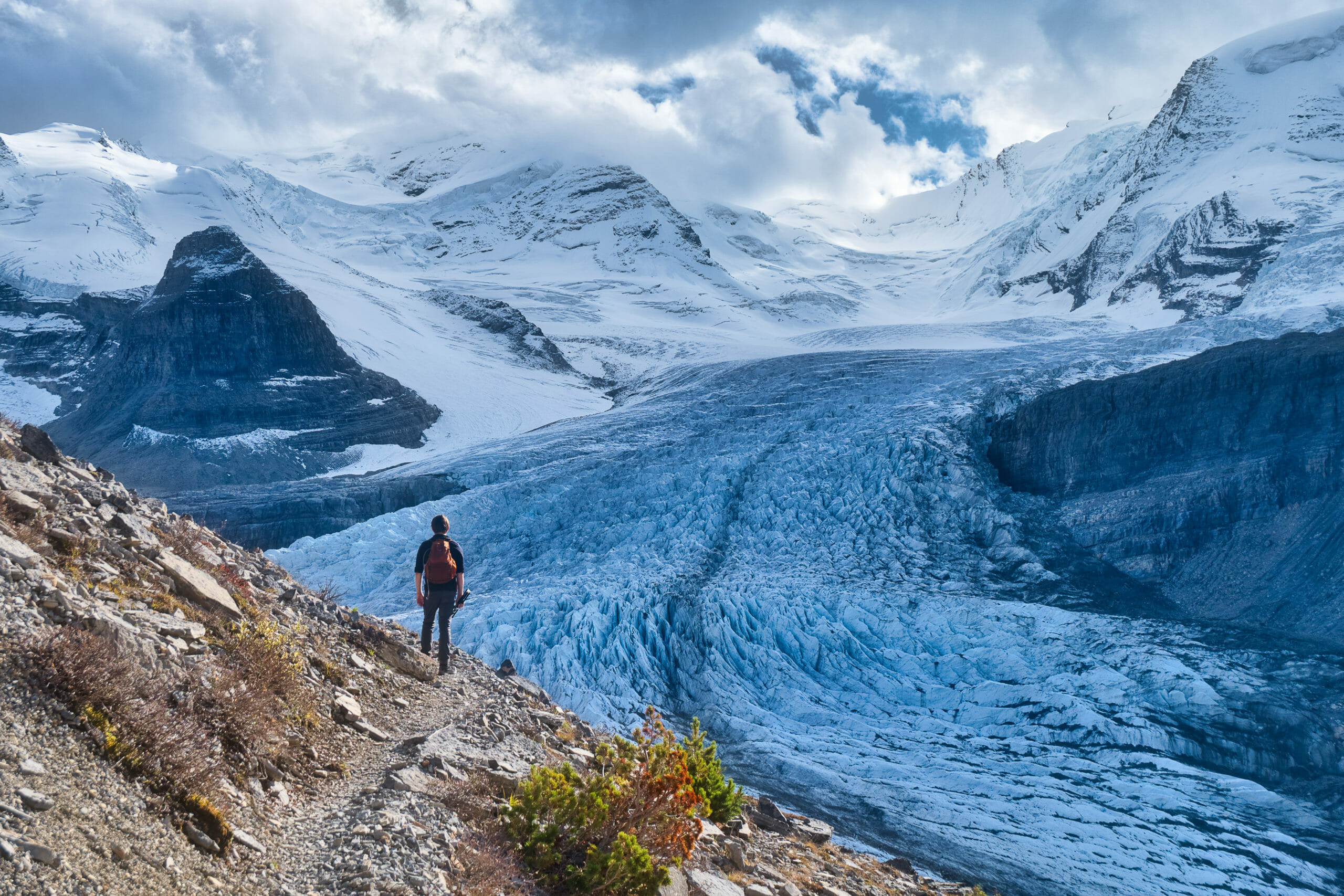 Male hiker walks around switchback in Snowbird Pass in front of Robson Glacier in Mount Robson Provincial Park on cloudy day