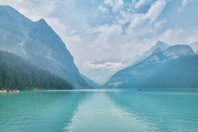 Sunlight shines a path down the middle of Lake Louise with mountains and glacier obscured by clouds and smoky wildfire skies