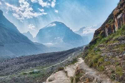 Sun rays break through clouds to shine down on mountains on Plain of Six Glaciers trail and striped cliffs on sunny smoky day