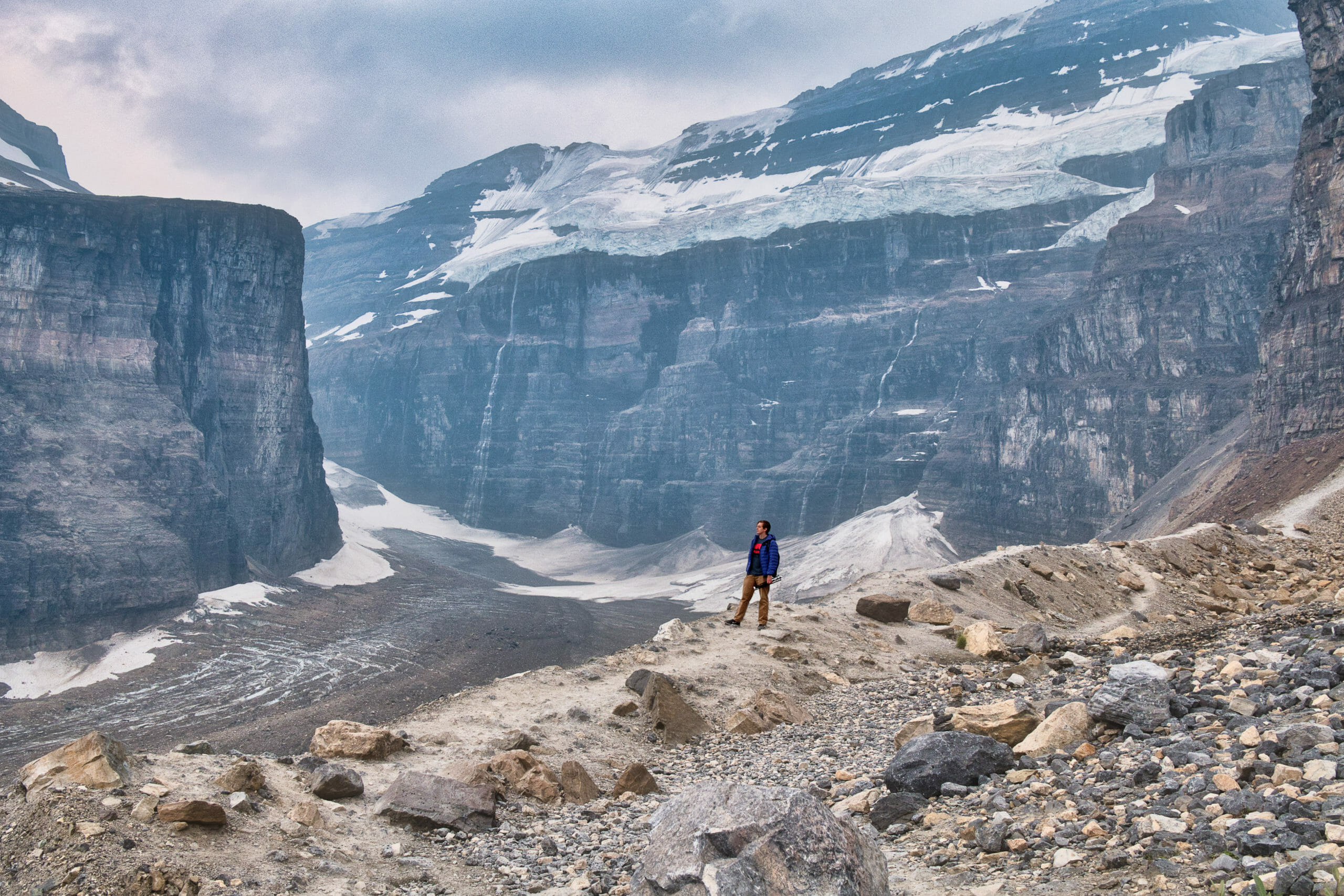 Male hiker on Lake Louise Plain of Six Glaciers trail surrounded by snow covered moraines under storm clouds and smoky skies